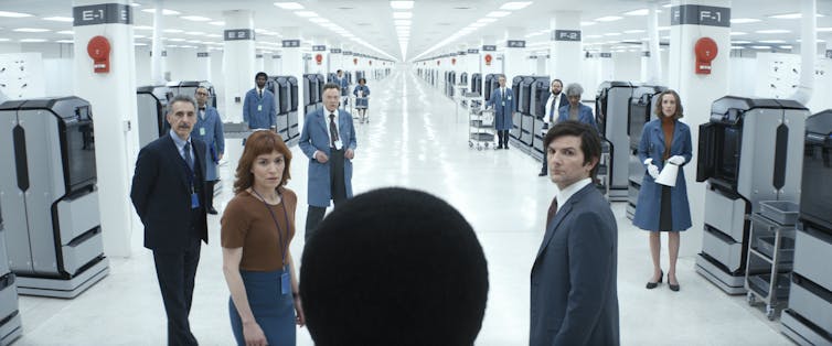 A group of people stand in a stark white room that has a long hallway with cubicles on the edge of the photo.