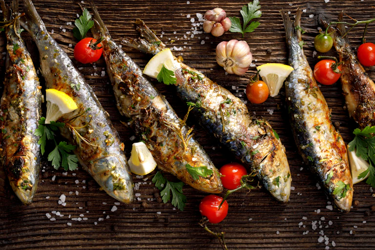 Fried sardines are places on a board with lemon wedges and tomato as garnish