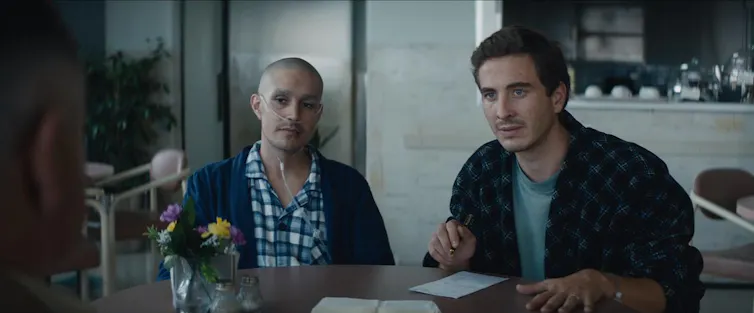 An ill man, bald and in pyjamas, sits at breakfast with a healthier looking man.