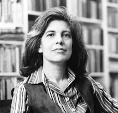 Grey-haired woman in front of bookselves, wearing a waistcoat over a striped shirt.