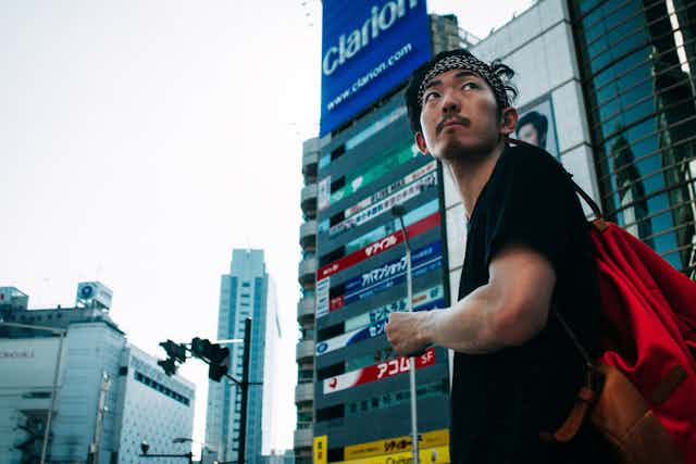 A man with a red backpack and bandana looks over his shoulder against a high-rise city backdrop.