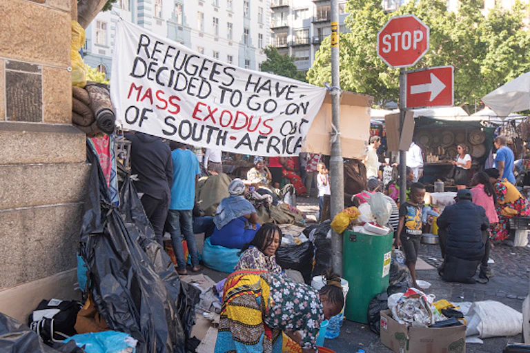 Men, women and children crowded on a pavement display a sign that says "Refugees have decided to go on a mass exodus out of Soiuth Africa'.