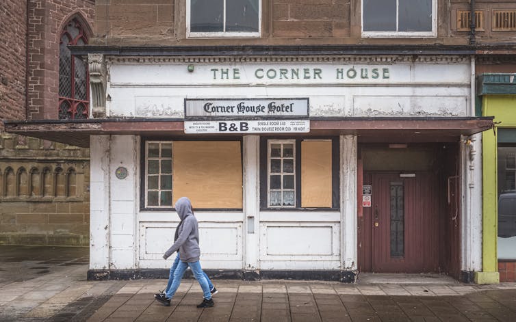 Two people in jeans walk past a boarded up B&B on a Scottish street.