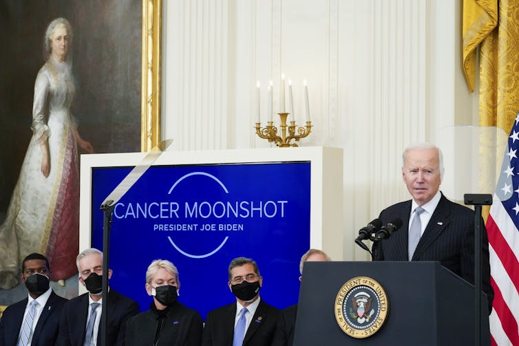 A man in a suit at a podium beside a blue display reading 'Cancer Moonshot'