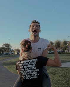 The back of a person's half-shaved head is seen and a t-shirt reading 'no homophobia, no violence, no racism, yes kindness yes love and this person is lifting up another person who is laughing joyously