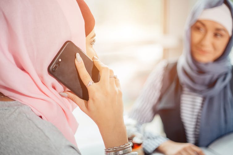 A woman in a pink headscarf talks on the phone as a woman in a blue headscarf looks on.
