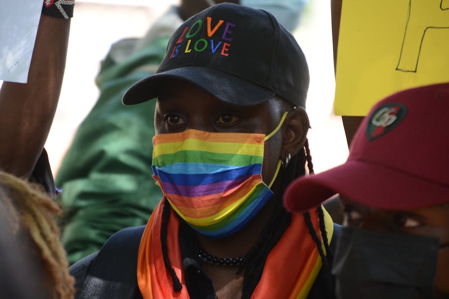 Close up of a protester surrounded by posters and people, a rainbow facemark on and a peak cap with the word 'love'.