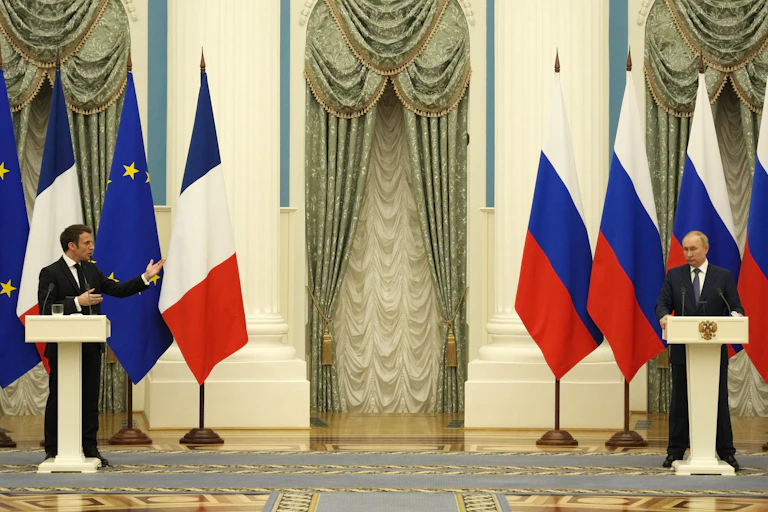 Two men stand far apart at podiums with Russian, EU and French flags behind them in an ornate hall.