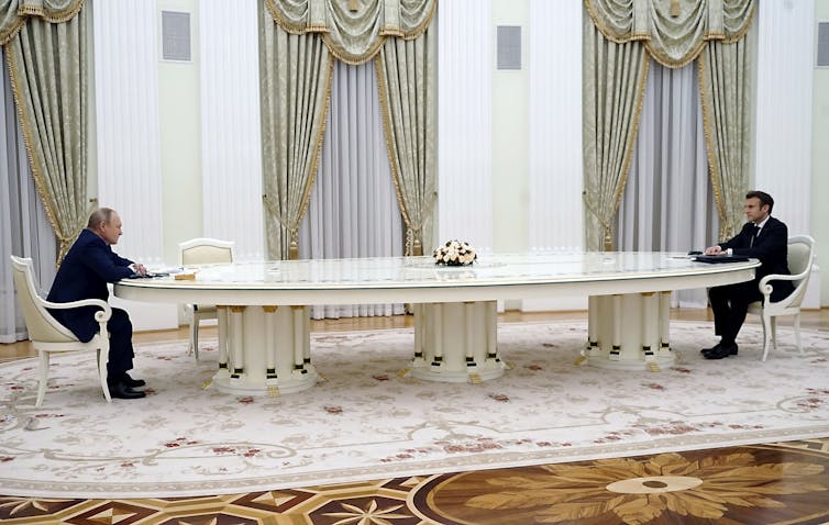 Two men sit far apart at opposite ends of a large, ornate white table in an ornate room with cream carpets and drapery