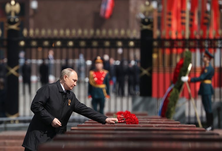 A balding man in a dark suit and overcoat lays a wreath on a casket.
