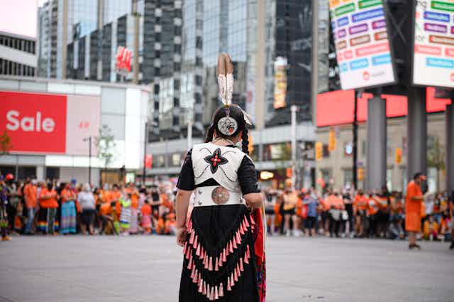 A young girl in braids wearing a jingle dress stands in front of a crowd at Yonge and Dundas