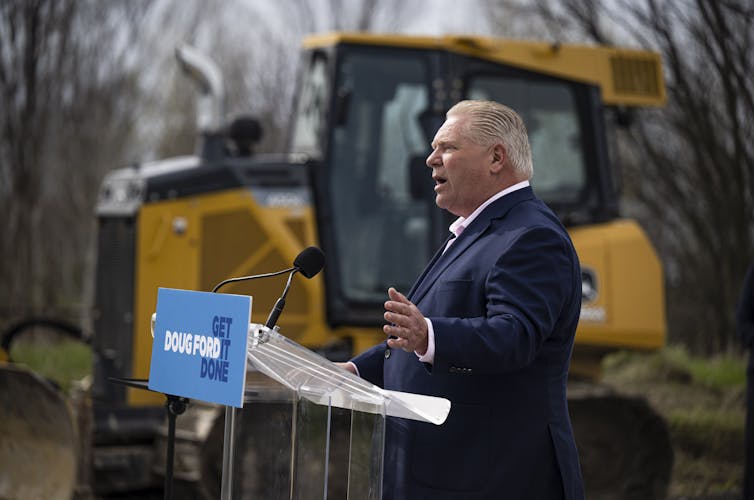 A man with blond-ish-grey hair in a navy suit speaks into a microphone, a large bulldozer in the background.
