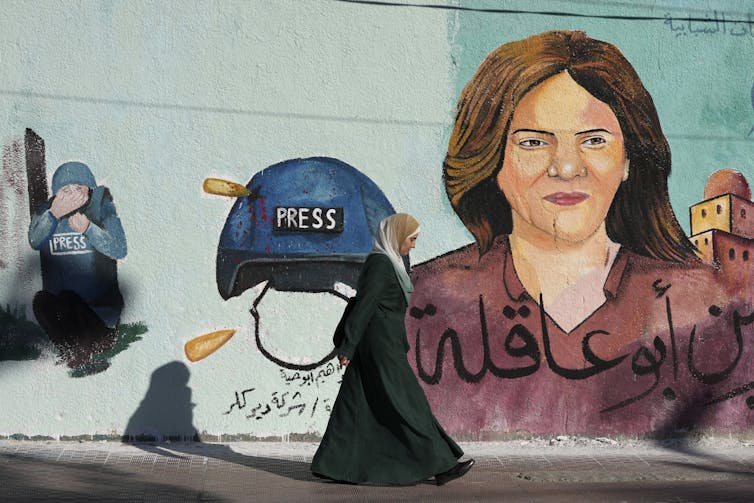 A women walks past a mural depicting slain journalist Shireen Abu Akleh and a helmet with 'PRESS' on it.