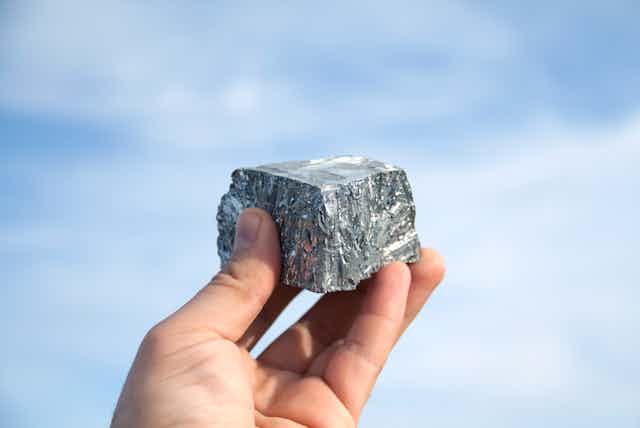 Hand holding block of silver zinc metal up against a backdrop of the sky