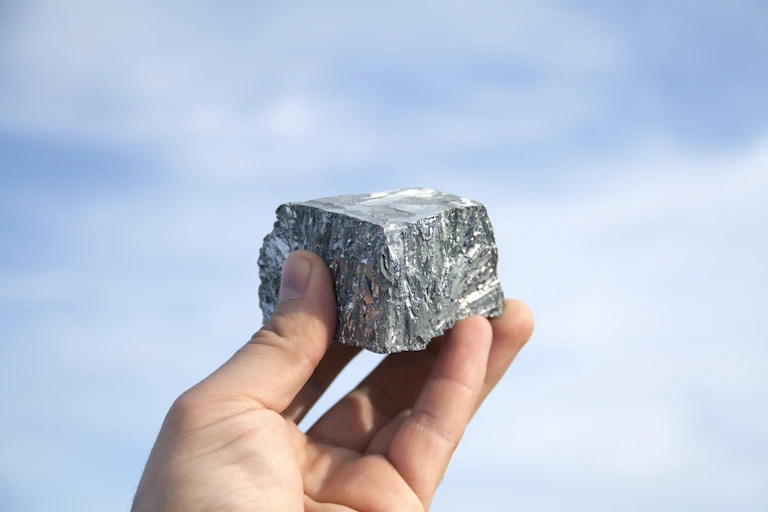 Hand holding block of silver zinc metal up against a backdrop of the sky