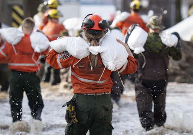 A group of people wearing orange jackets and black pants wade through shin-deep water, carrying white bags on their shoulders. 