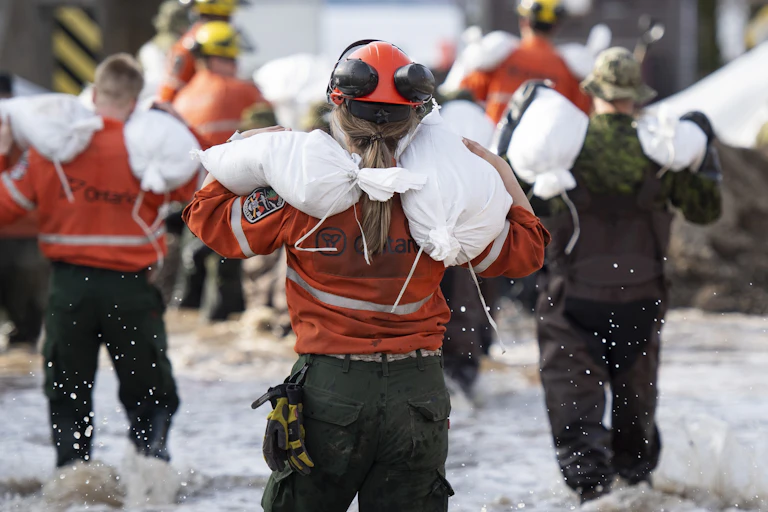 A group of people wearing orange jackets and black pants wade through shin-deep water, carrying white bags on their shoulders.