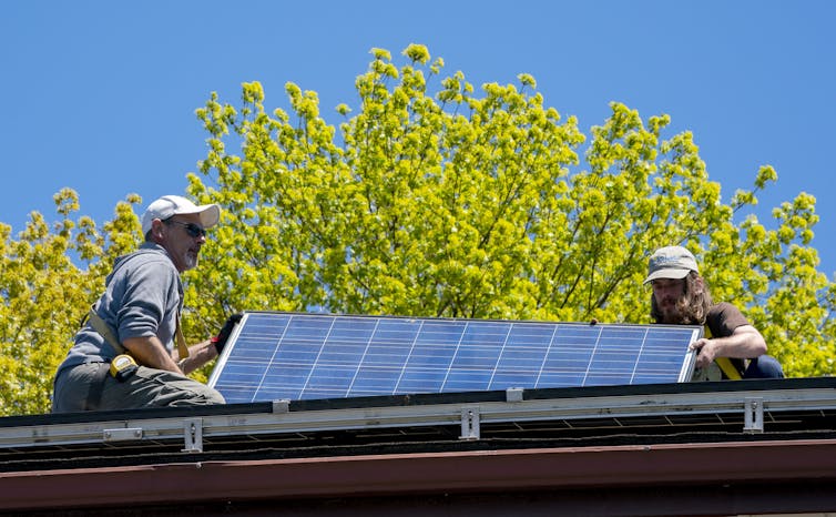 Two men wearing baseball caps position a solar panel on a roof.