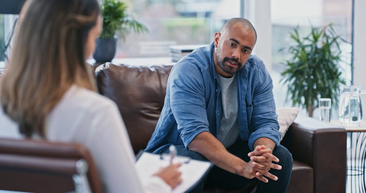 Person with hands clasped talking to therapist writing on a clipboard.