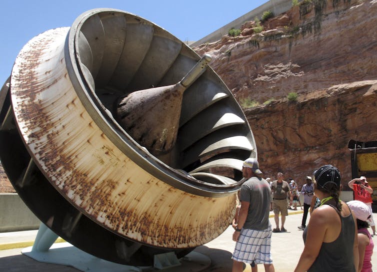 People look at a partially rusting turbine set up for display outside. It's about twice the height of the tallest person in the crowd.