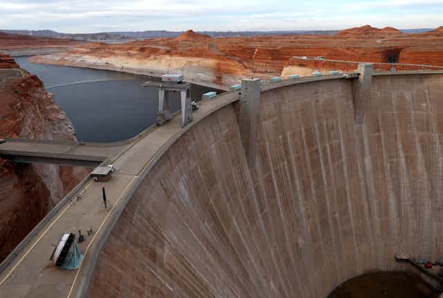 A tall, cement dam curves across the canyon with the reservoir behind it. The bottom half of the walls of the reservoir are white where they were once covered with water.