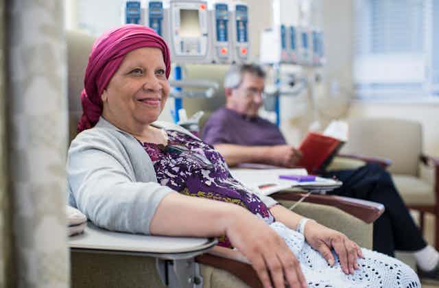 A smiling woman in a bright pink turban sits with an IV tube attached to her wrist. In the background, a man with an IV sits in another chair, reading.