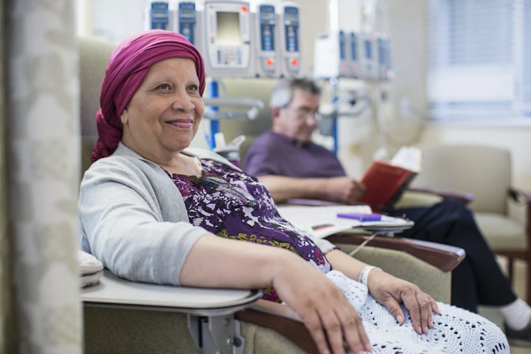 A smiling woman in a bright pink turban sits with an IV tube attached to her wrist. In the background, a man with an IV sits in another chair, reading.
