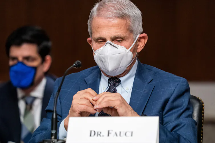 white-haired man in jacket and tie seated at mic with 'Dr. Fauci' on name plate