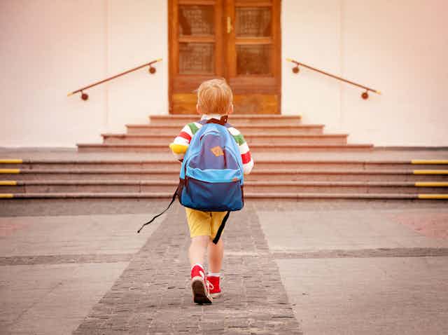 A small boy in yellow shorts and red trainers carries a big blue rucksack on his back as he walks towards a school door.