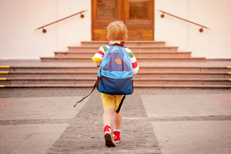 A small boy in yellow shorts and red trainers carries a big blue rucksack on his back as he walks towards a school door.