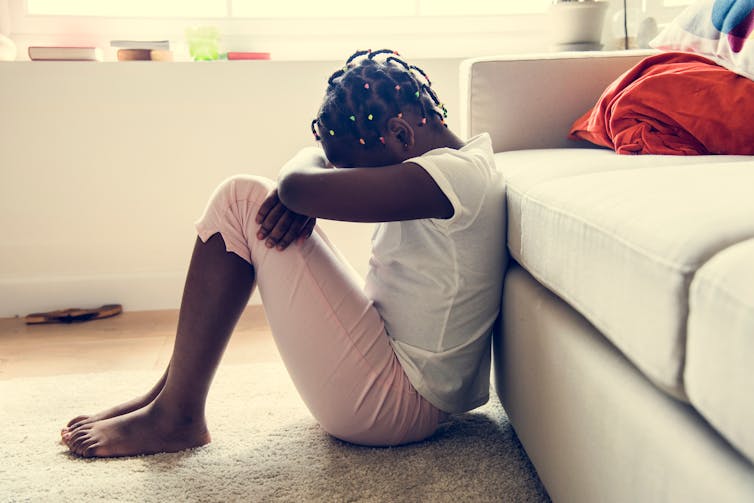 A young black girl rests her head on her forearms on her knees, sitting on the floor, barefoot, in a living room setting.