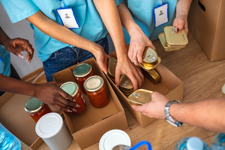 Hands of volunteers sorting jars of pasta sauce and tins of fish into carboard boxes