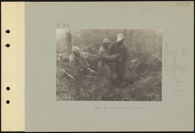 Black and white image of three male soldiers, one injured, on a battlefield with trees and mud.