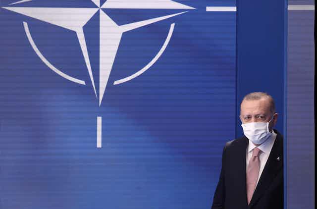 Turkey's President Recep Tayyip Erdogan wearing a suit and a face mask walks onto the stage with the NATO symbol behind him.