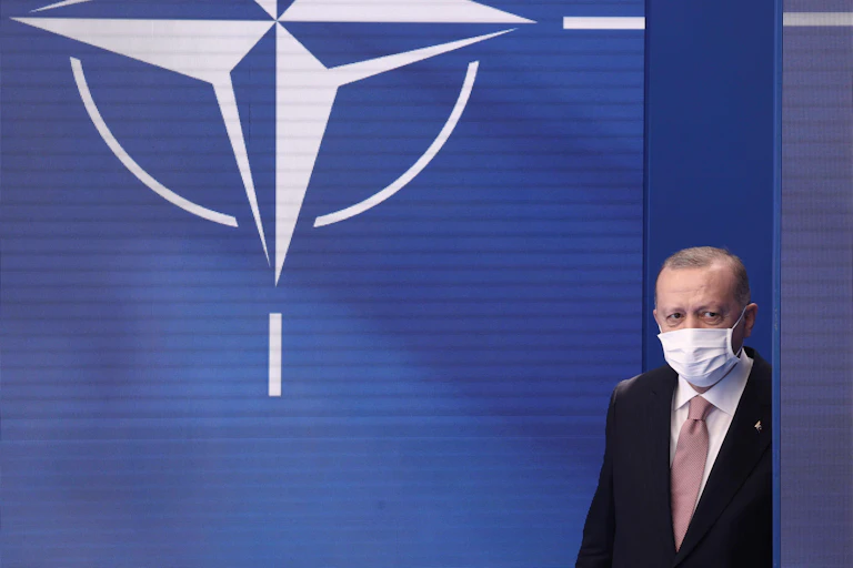 Turkey's President Recep Tayyip Erdogan wearing a suit and a face mask walks onto the stage with the NATO symbol behind him.