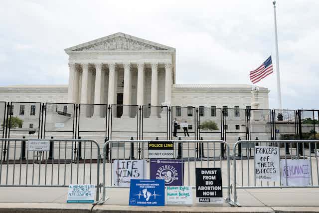 The Supreme Court building is shown behind two barriers of fencing, with signs like 'life wins' and 'divest from big abortion now!' outside of the fence