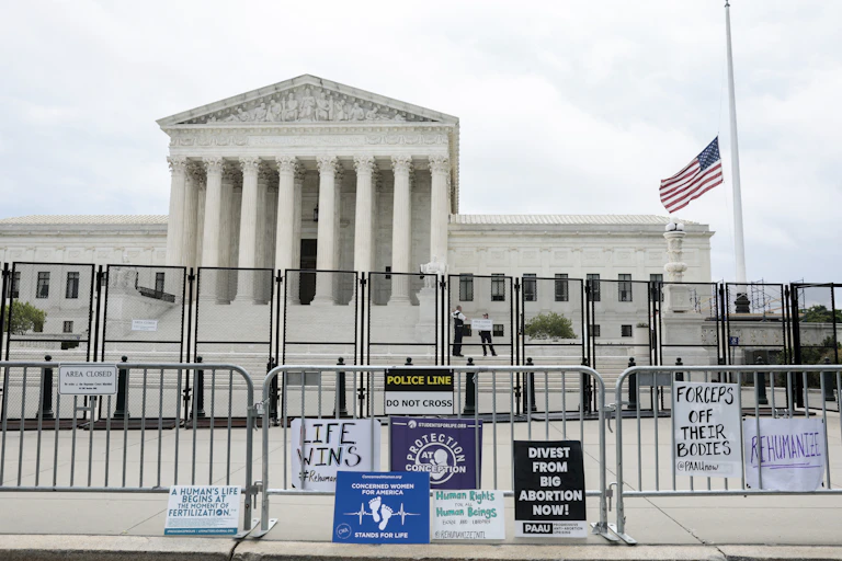 The Supreme Court building is shown behind two barriers of fencing, with signs like 'life wins' and 'divest from big abortion now!' outside of the fence