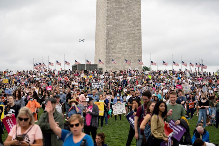 A crowded field of people outside the Washington Monument, surrounded by American flags, hold signs that say things like 