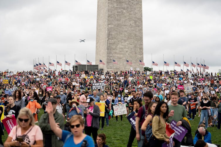A crowded field of people outside the Washington Monument, surrounded by American flags, hold signs that say things like