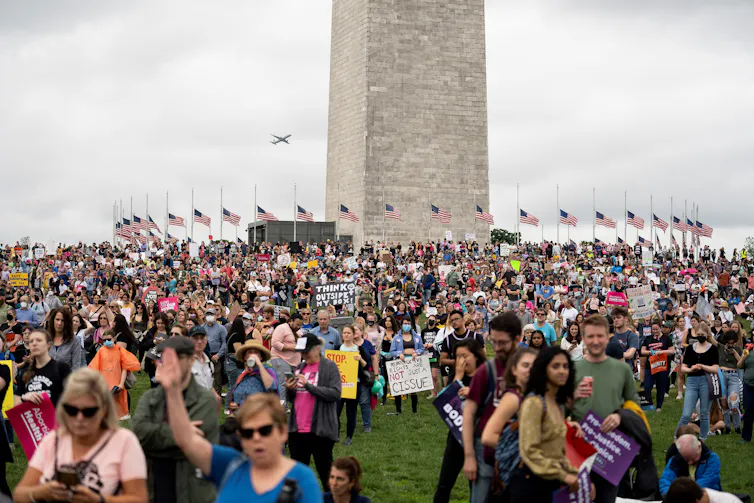 A crowded field of people outside the Washington Monument, surrounded by American flags, hold signs that say things like