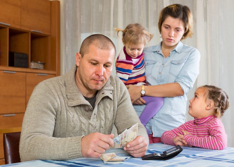 A man sitting at a table looking at mail, with a child beside him, and a woman behind him holding another child.