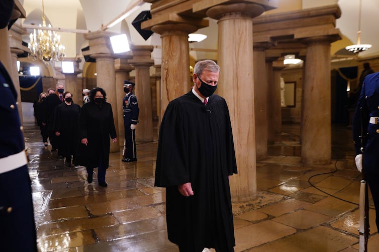 Chief Justice Roberts wears a black robe and mask and walks down a government building hall, with Sonia Sotomayor, Elena Kagan and other justices behind him.