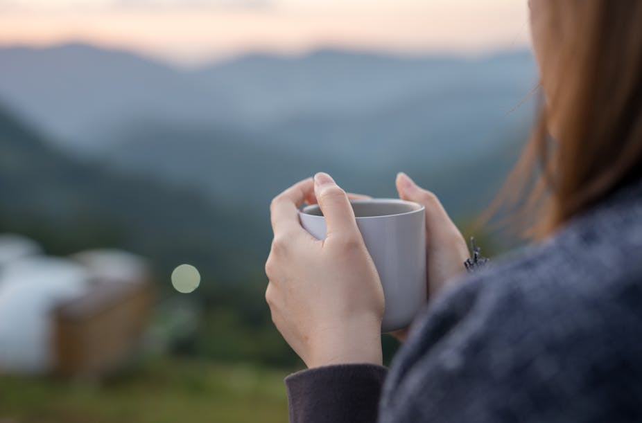 A woman with long hair cradles a mug and looks over a mountain view