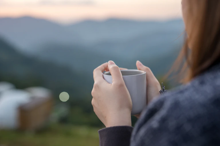 A woman with long hair cradles a mug and looks over a mountain view