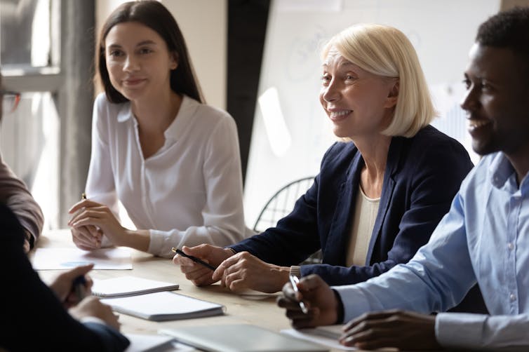 An older blonde woman sits between two younger people in an office meeting.