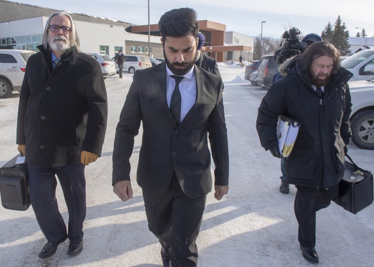 A man wearing a suit walks with his head down in a snow-covered parking lot with two people flanking him