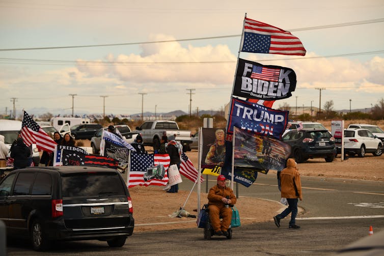At a gathering of people and trucks, one person holds flags with a swear word statement against Biden and in favor of Trump.