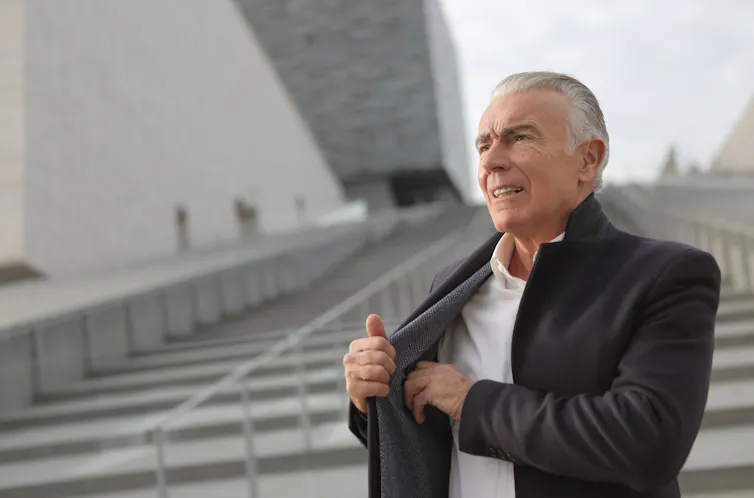 A grey-haired man walks down a stone staircase tucking his phone into an inside jacket pocket.