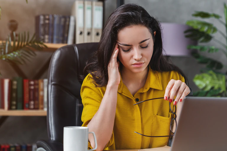 Woman sitting in front of open laptop pressing a hand to her temple and closing her eyes