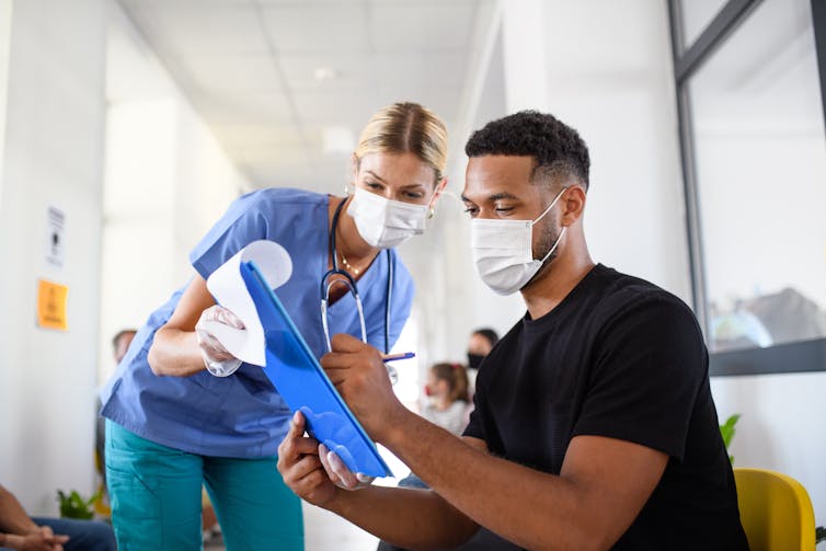 A nurse assists a young man to fill out a form. Both are wearing masks.